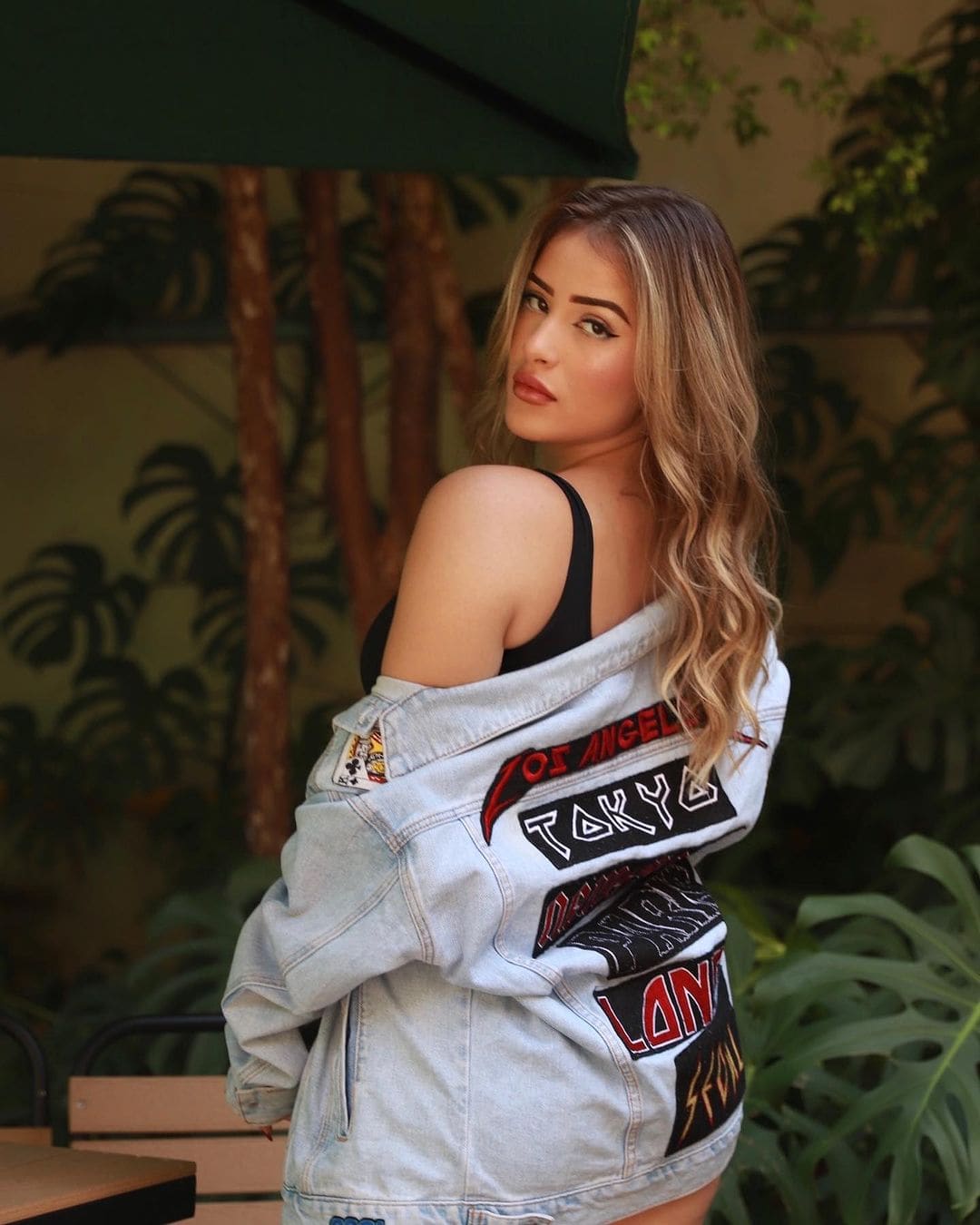 Looking back over her shoulder, a woman in a black tank top and patched denim jacket stands among tropical plants.