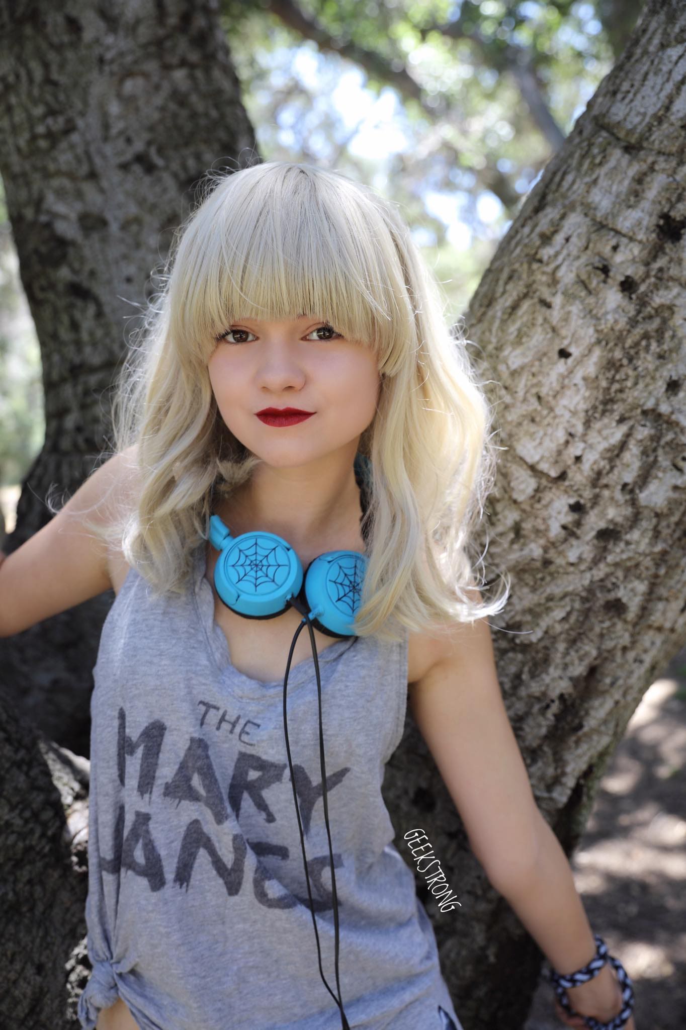 A young blonde woman with bangs stands between tree trunks in a grey sleeveless top, headphones around her neck, hand on the bark.