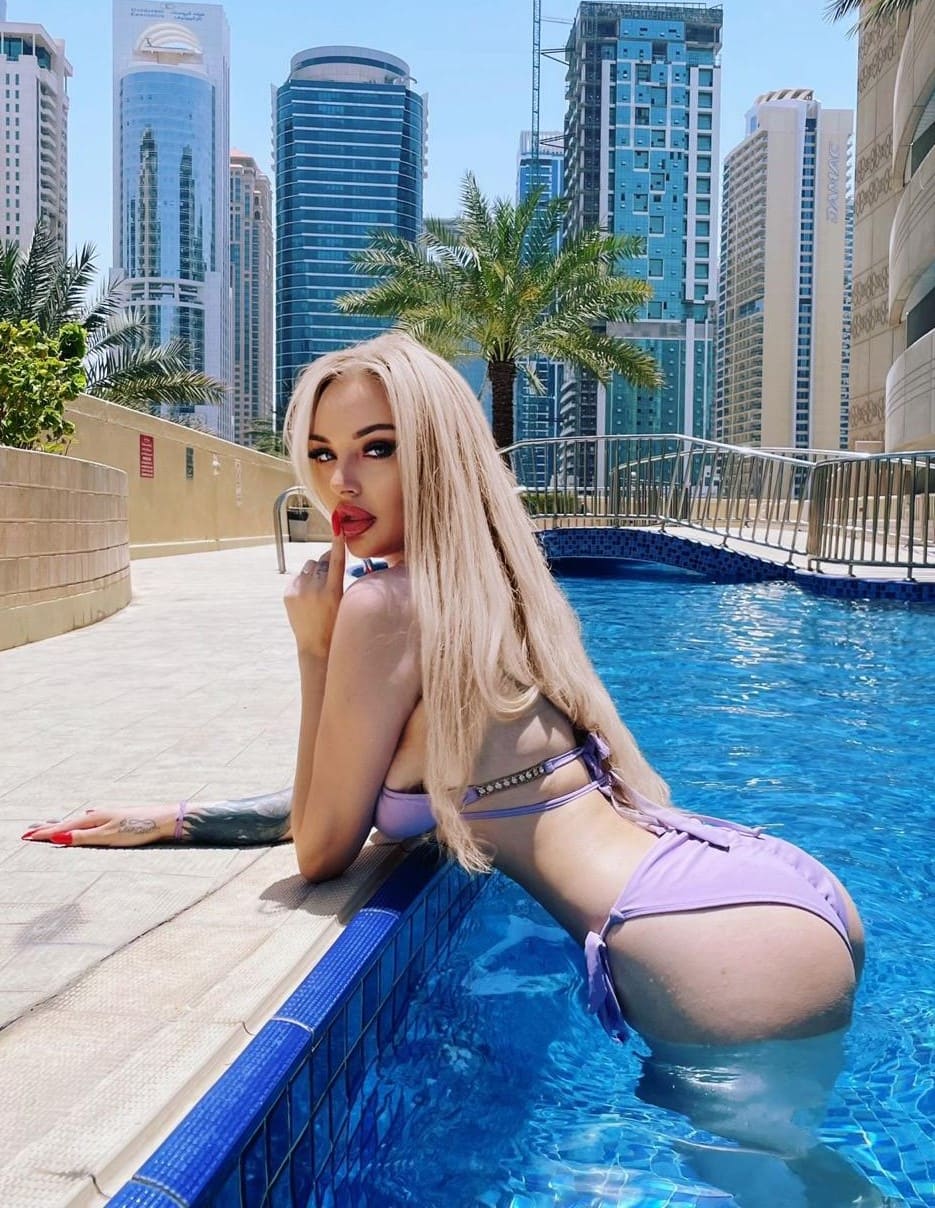 A woman in a light purple bikini leans on the edge of a blue-tiled pool, with a city skyline and palm trees visible behind her.