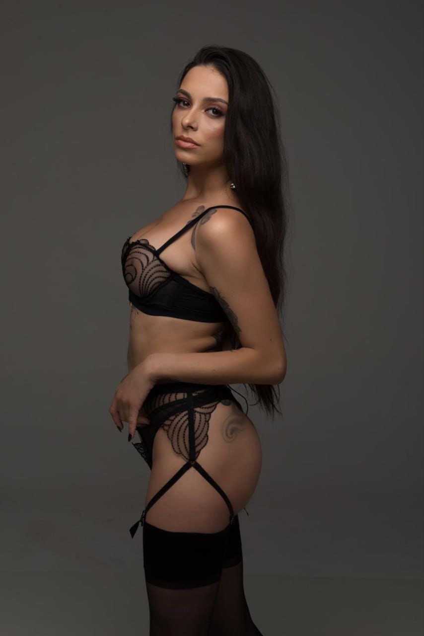 A woman stands in profile against a gray backdrop, wearing a black lace bra, briefs with garter straps, and sheer thigh-high stockings, showing tattoos.