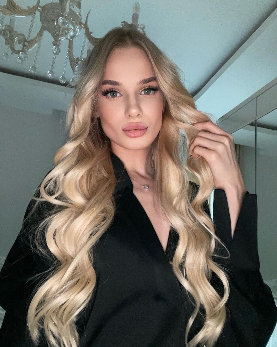 Rosalie stands indoors by a chandelier in a black V-neck shirt, touching her wavy blonde hair.