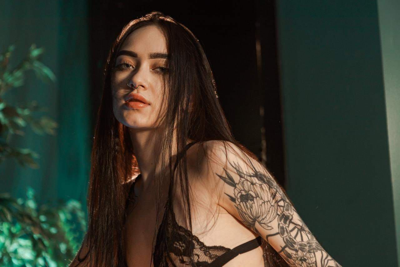 Vilena in a black lace bralette and floral arm tattoos poses against a dim teal wall with a plant nearby.