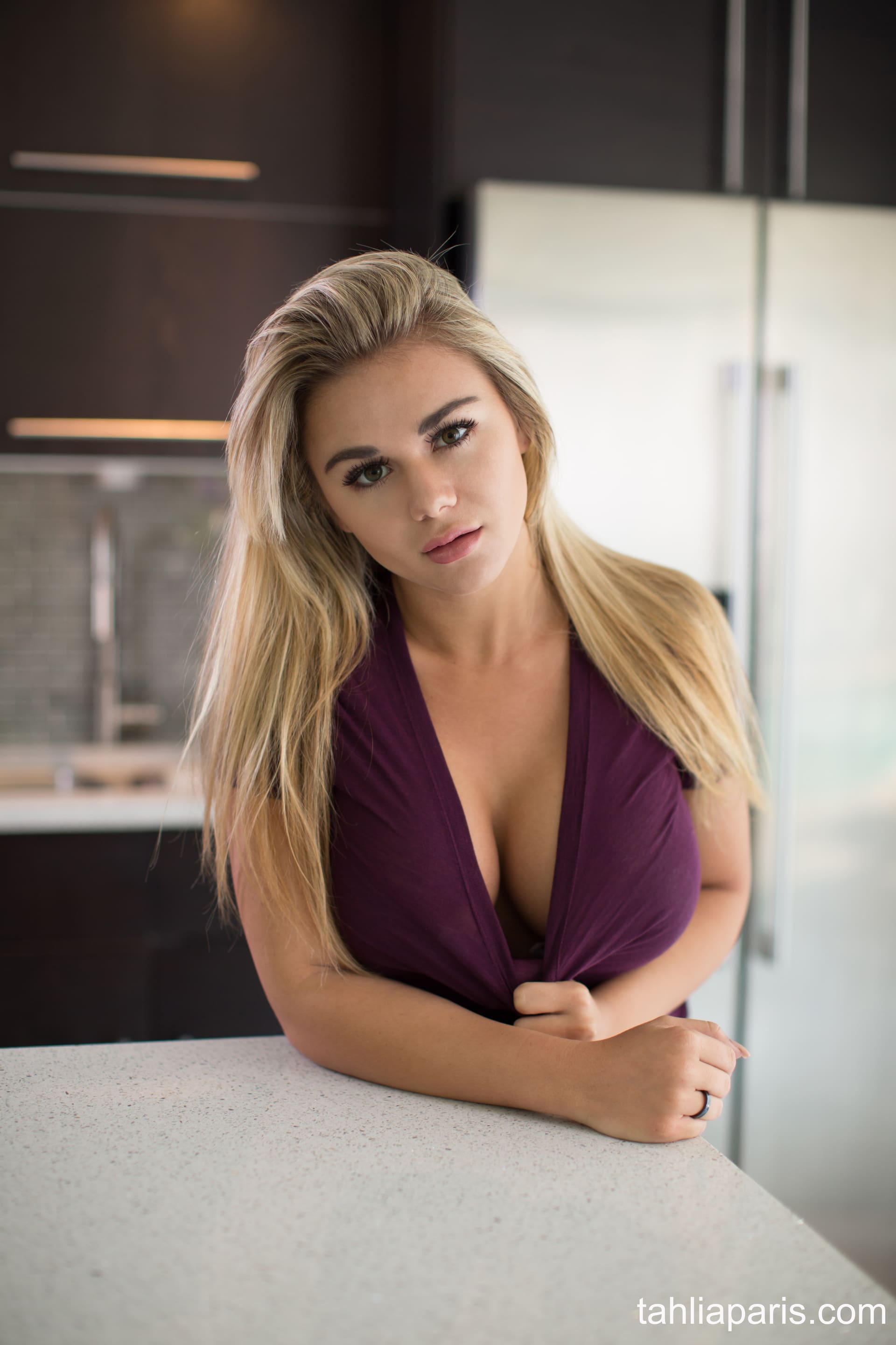A blonde woman leans on a white counter in a dark kitchen, wearing a purple wrap top with a plunging neckline and a steel fridge behind.