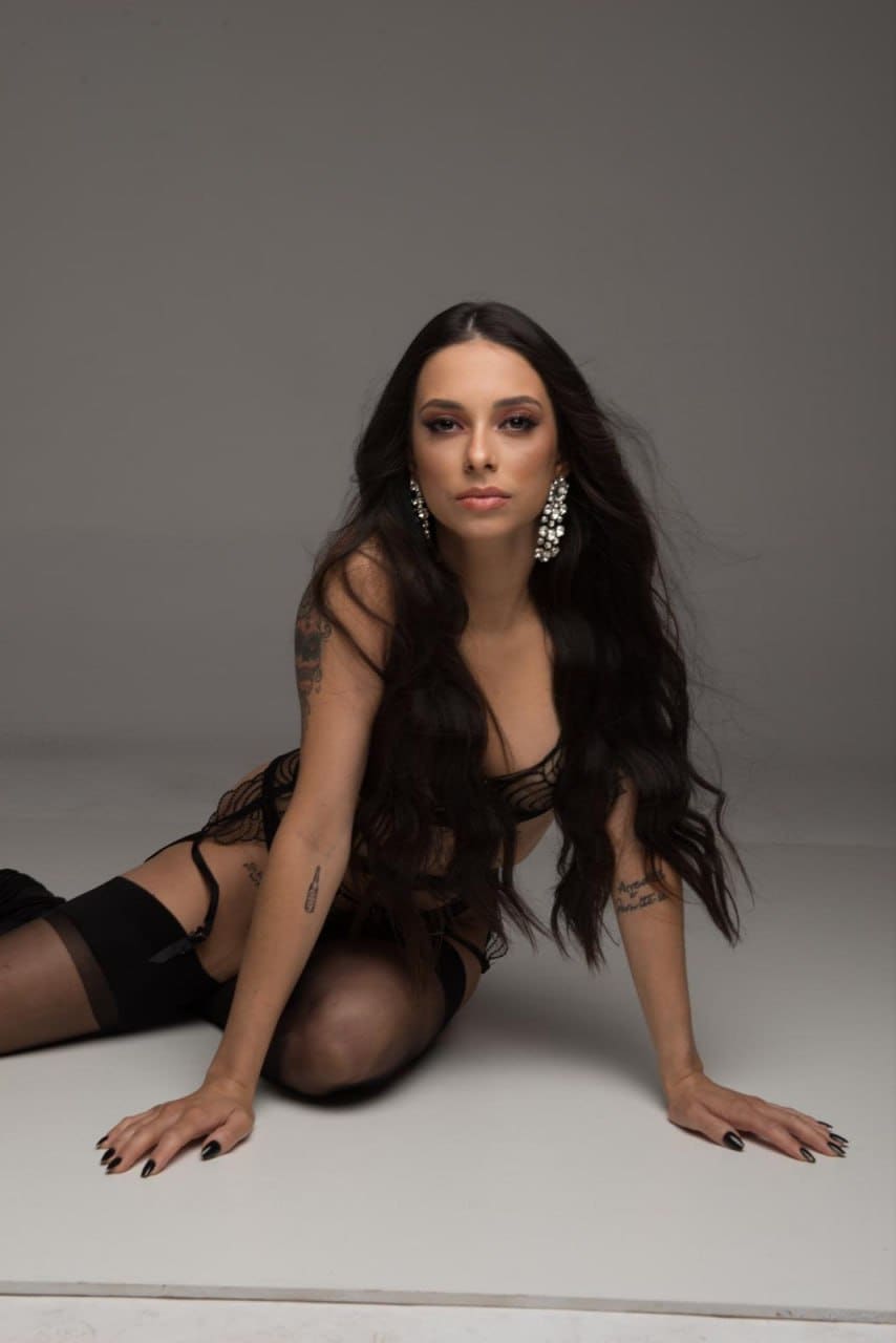 A woman with arm tattoos kneels on a white studio floor, wearing a black lace bra and gartered stockings, with large dangling earrings and dark nails.