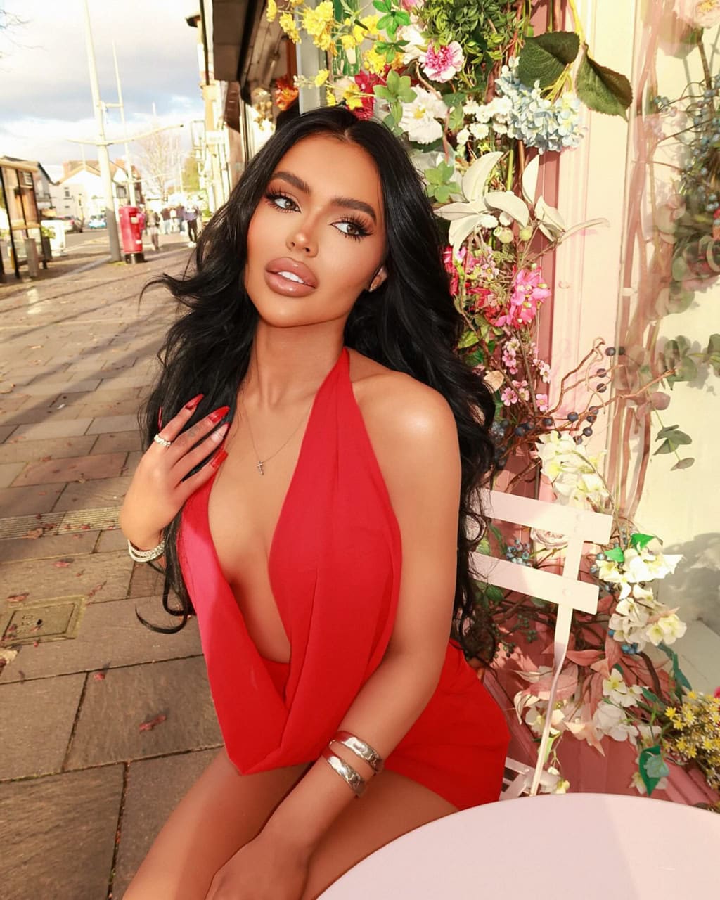 Nino sits at an outdoor table in a red halter dress, hand near chest, against a flower-decorated wall and street.