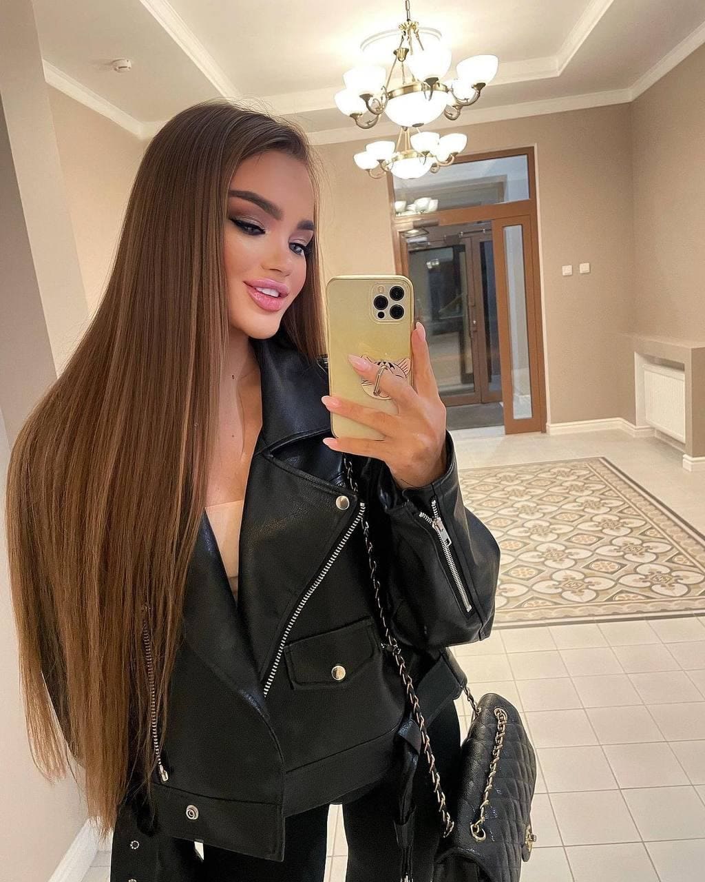 Karina takes a mirror selfie in a tiled hallway, wearing an open black leather jacket over a light top and a quilted shoulder bag.