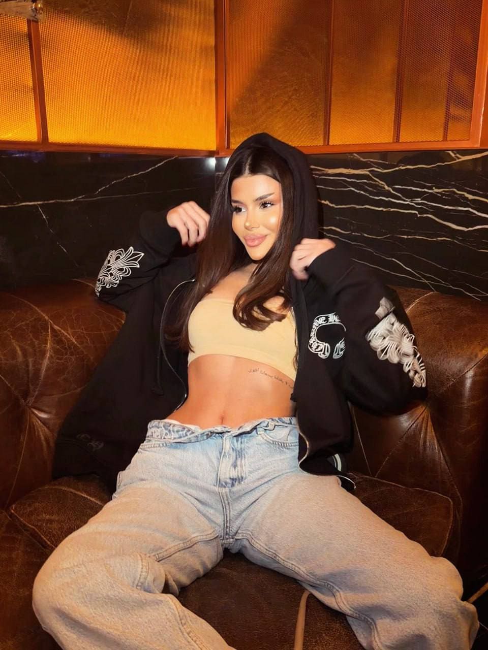 Seated on a brown leather couch in a black embroidered jacket, beige crop top, and baggy jeans.