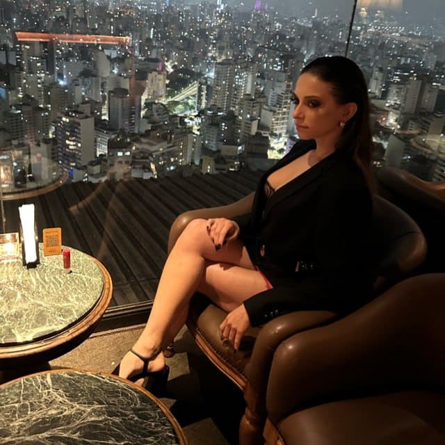 A woman in a black blazer and strappy heels sits in a brown leather armchair, with a nighttime city skyline visible through the window behind her.