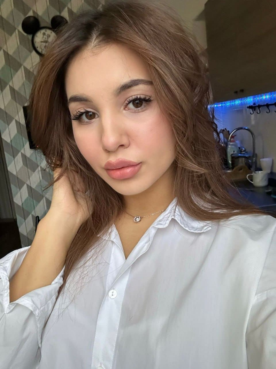 Tessa wears a white button-up shirt and thin necklace, posing with a hand near her neck against a patterned wall and kitchen counter.