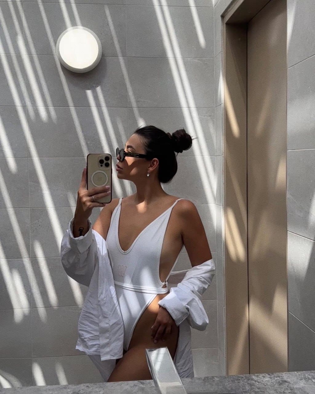 A woman with dark hair in a bun and sunglasses takes a mirror selfie in a white deep-V one-piece swimsuit and open shirt, standing in a tiled room with striped shadows.