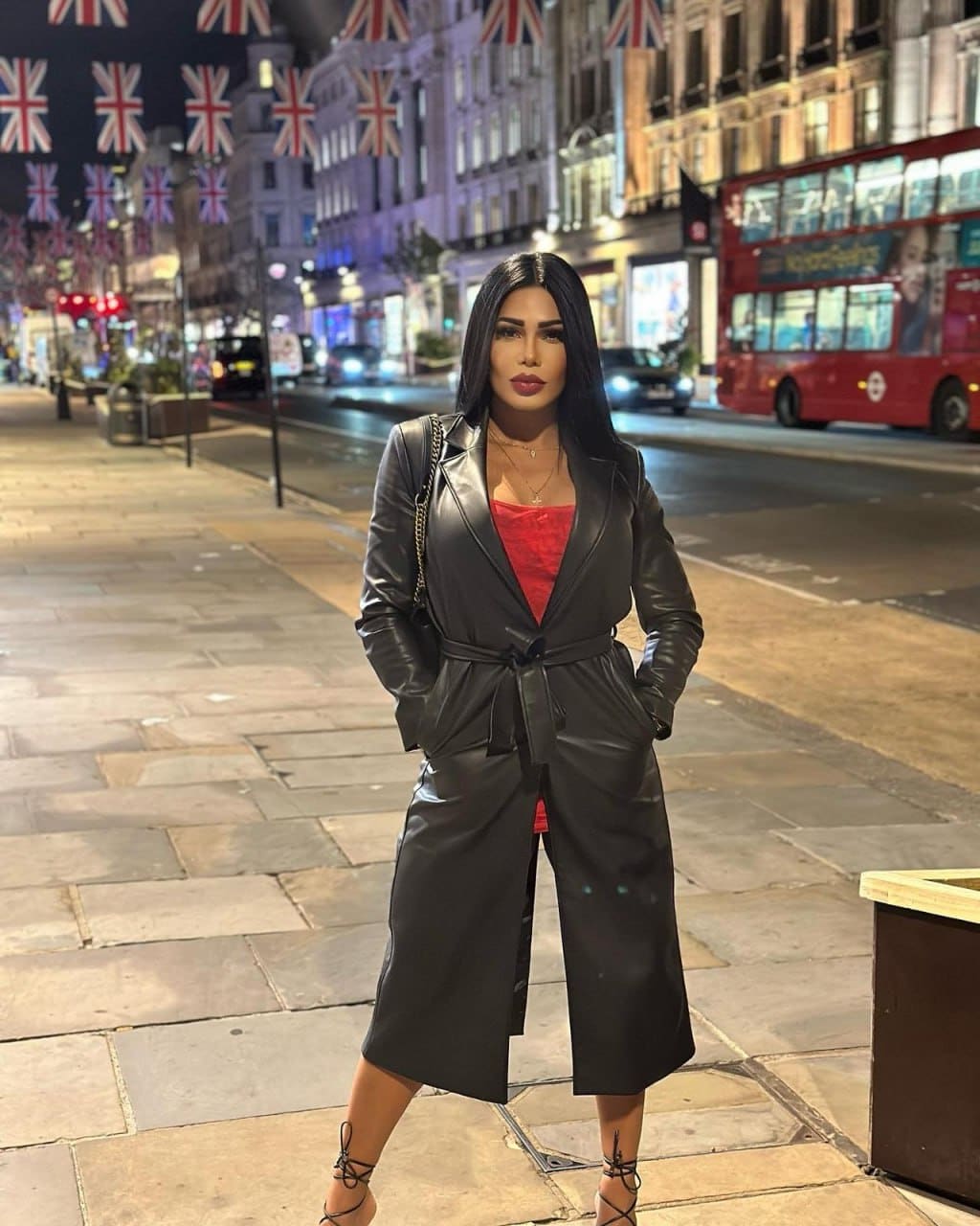 A woman in a black leather trench coat stands on a night sidewalk, with a red double-decker bus and Union Jack flags in the background.