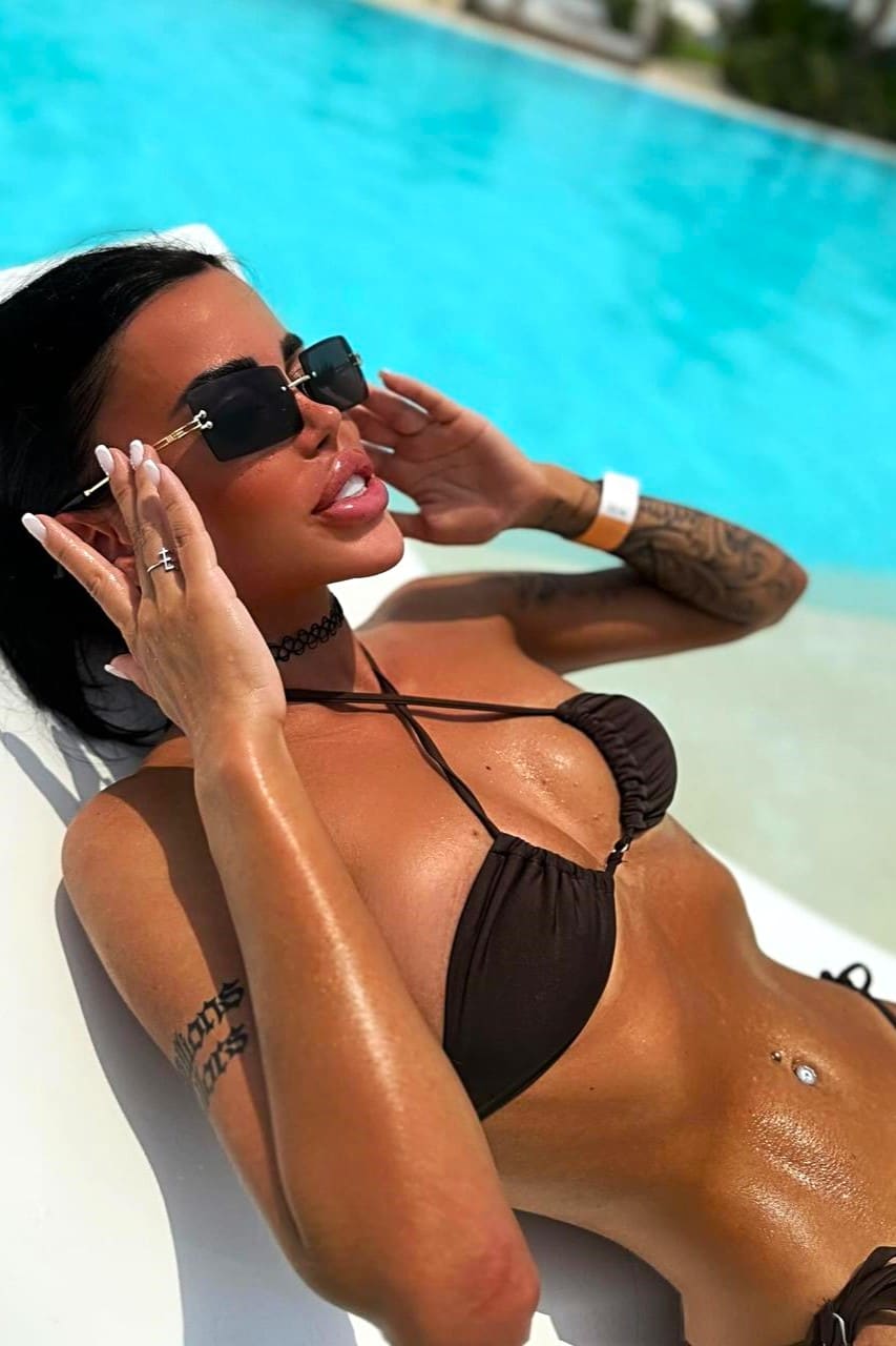 A woman lies on a white lounge chair by a blue pool in a dark bikini top and sunglasses, hands near her face, wearing a choker.