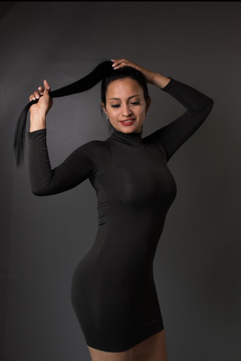 Davina stands in a studio against a gray backdrop, wearing a black turtleneck mini dress and holding her long dark hair up with both hands.