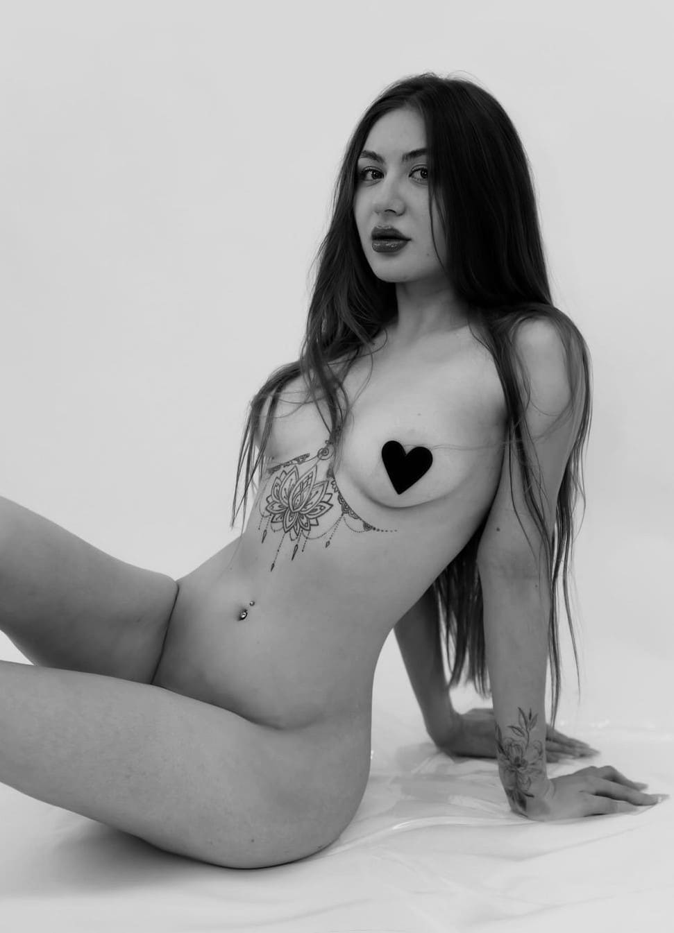 Black-and-white portrait of a woman seated on a white sheet, leaning back with legs bent, showing a torso tattoo and piercing with a heart graphic.