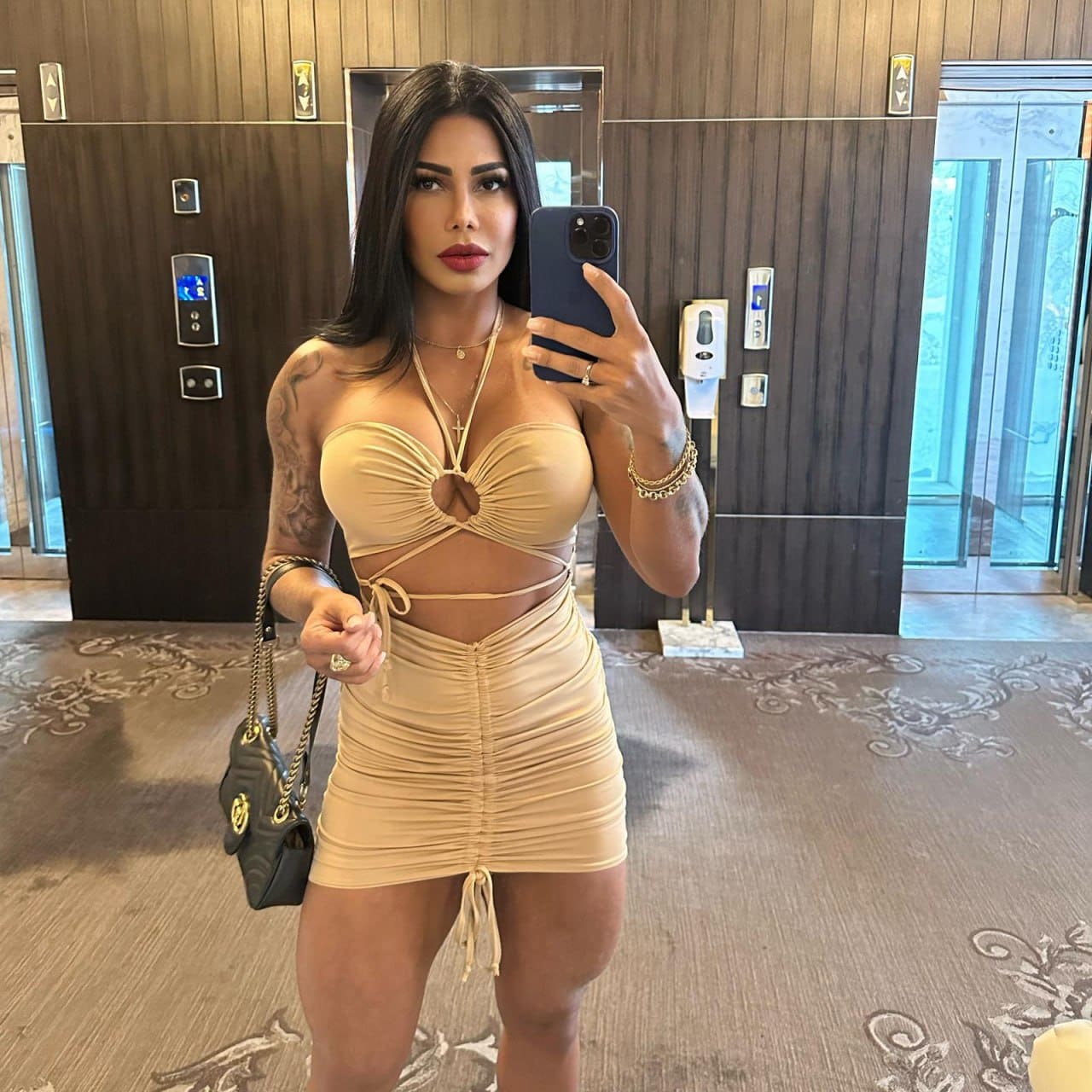 Jully takes a mirror selfie in a hotel lobby, wearing a beige cut-out top and ruched mini skirt while holding a phone and handbag.