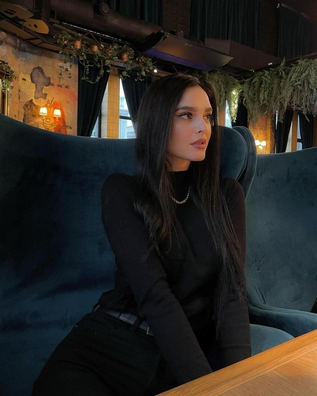 A woman in a black turtleneck and pants sits in a teal velvet chair in a dim room with plants and sconces.