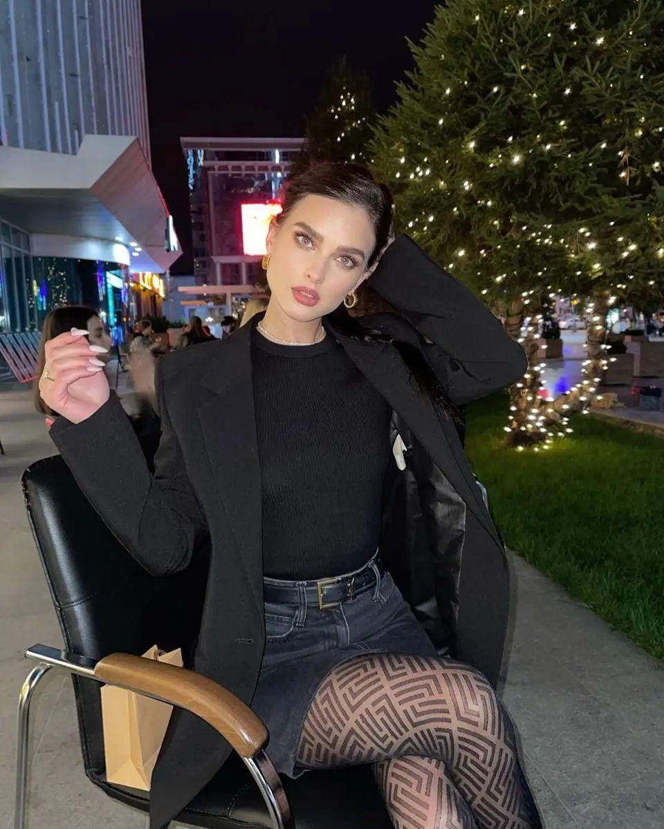 A woman in a blazer and sheer tights sits on a chair at night, with a lit plaza and trees behind her.