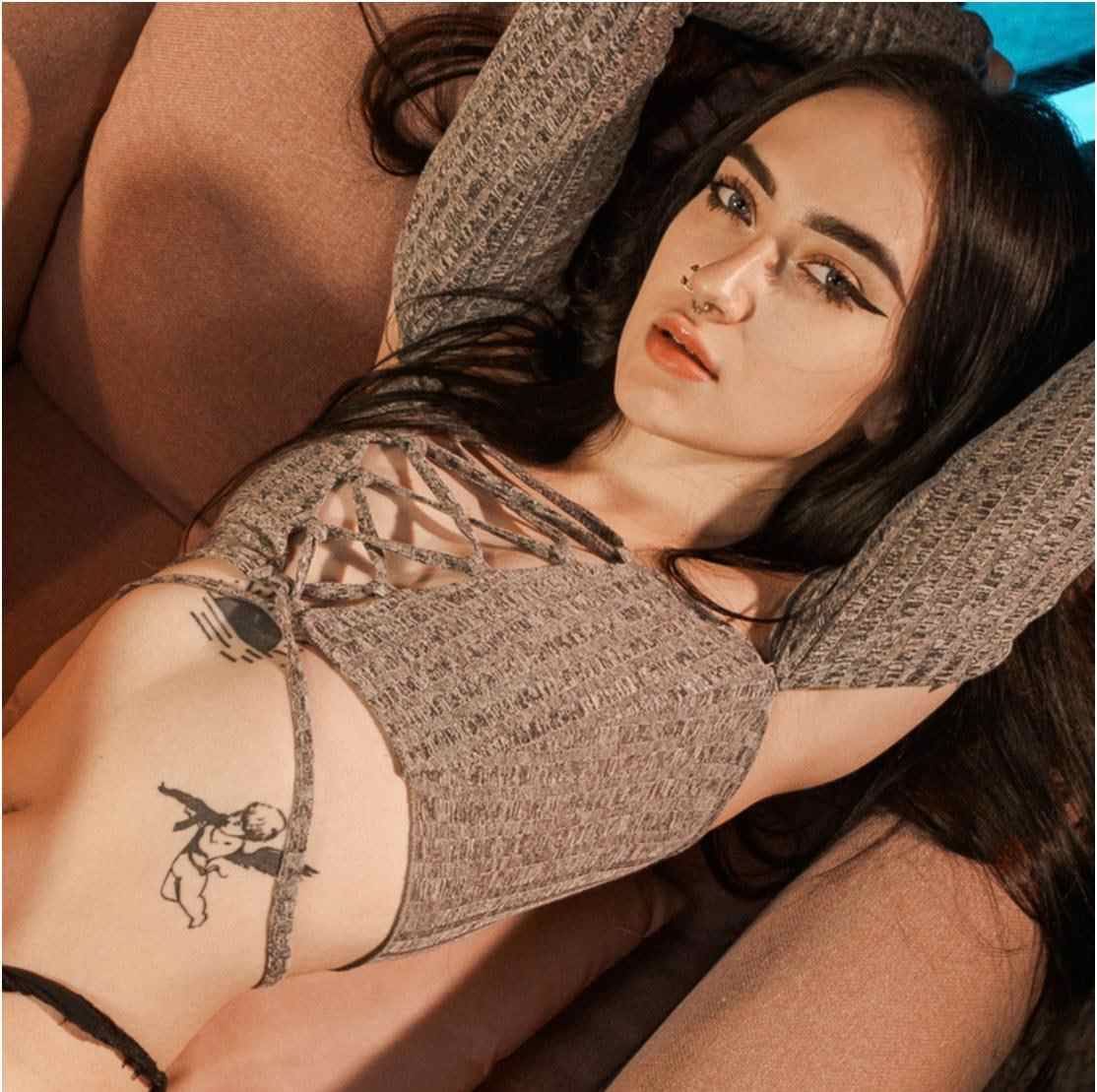 A woman in a grey knit crop top lies back with arms behind her head, revealing a side tattoo against a brown cushion.