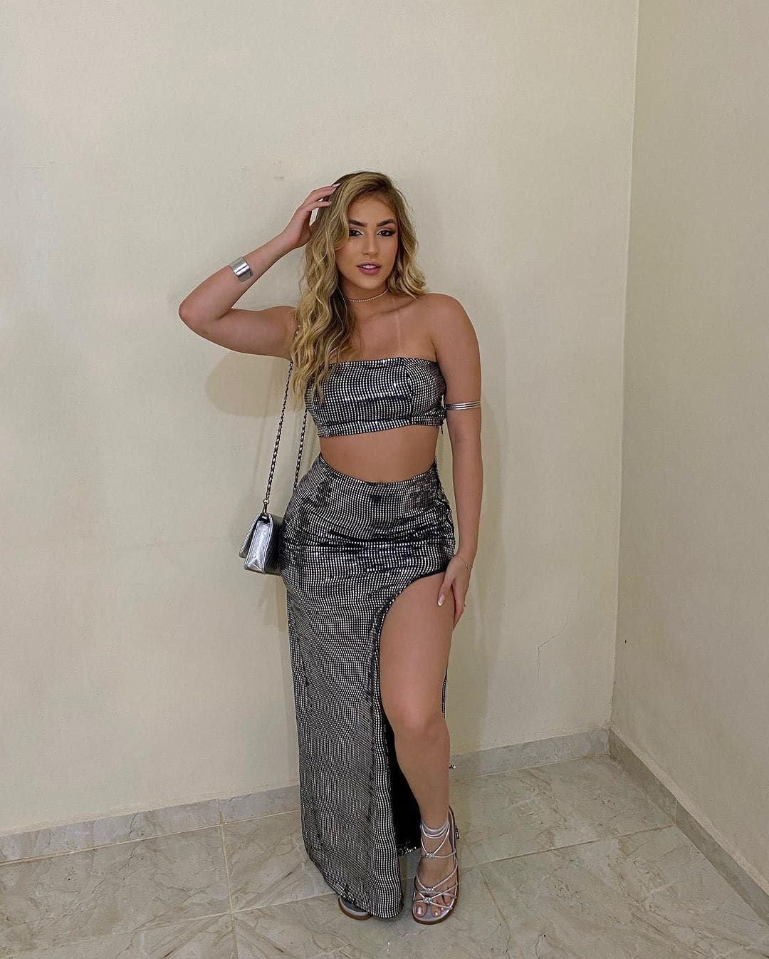 Standing on a tiled floor against beige walls, a woman wears a silver sequined crop top and high-slit skirt with a chain bag.