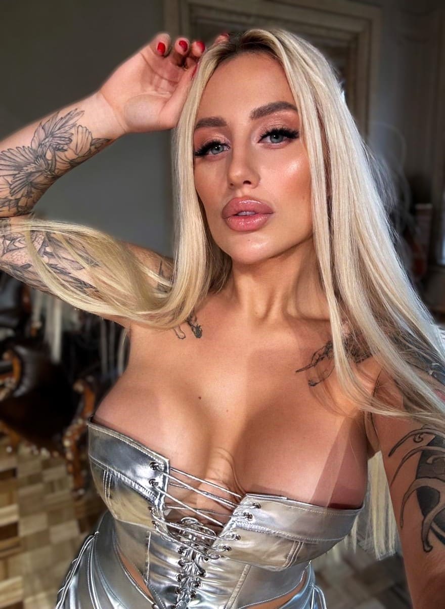 Gretta with blonde hair and tattoos wears a silver lace-up corset, posing indoors with her right hand raised to her head.