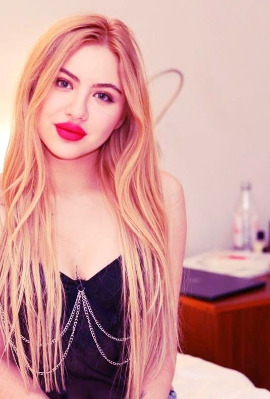 A blonde woman with pink lipstick sits facing the camera in a black strapless top with silver chains, in a room with a wooden desk and laptop.