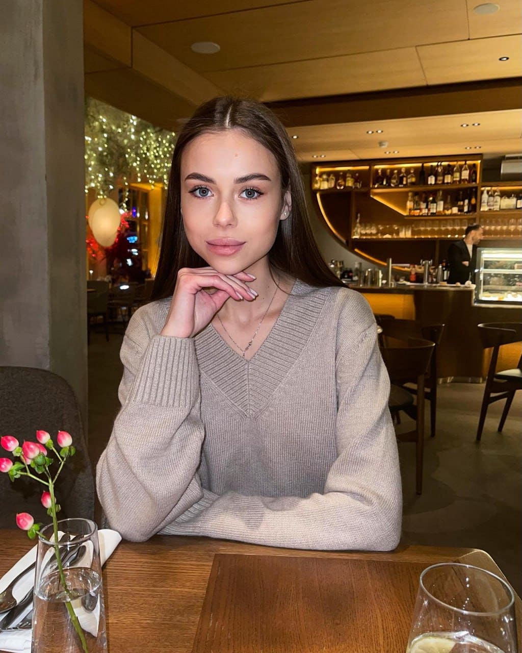 Janelle sits at a wooden restaurant table in a beige V-neck sweater, chin resting on her hand, with flowers and bottles behind.