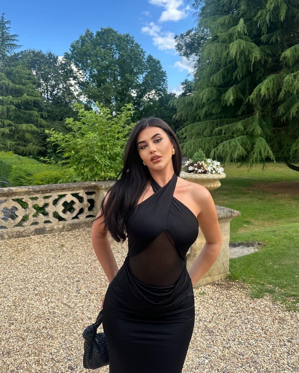 A woman stands on a gravel path before a stone balustrade in a black halter dress with a sheer midriff panel, holding a small black bag.