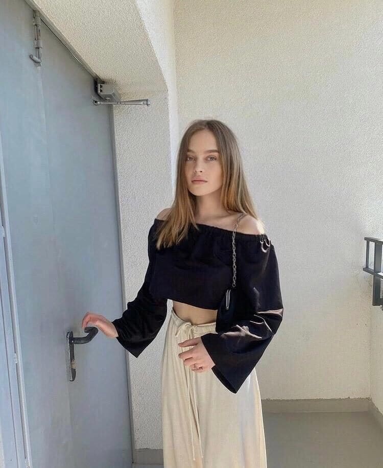 Bella stands on a balcony in a black off-the-shoulder crop top and cream skirt, holding a grey door handle against a white stucco wall.