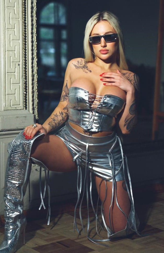 Wearing sunglasses and a silver corset with fringed skirt, a tattooed blonde kneels on a wooden floor near a stone column.