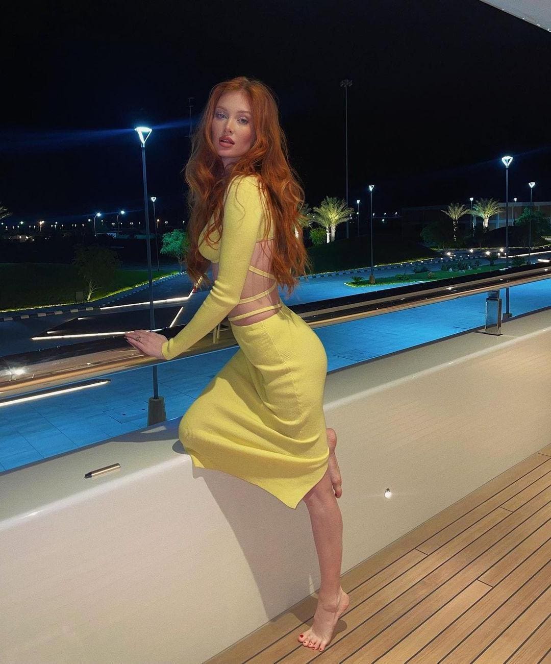 She sits by a pool at night in a yellow dress with a cutout back, one leg bent.
