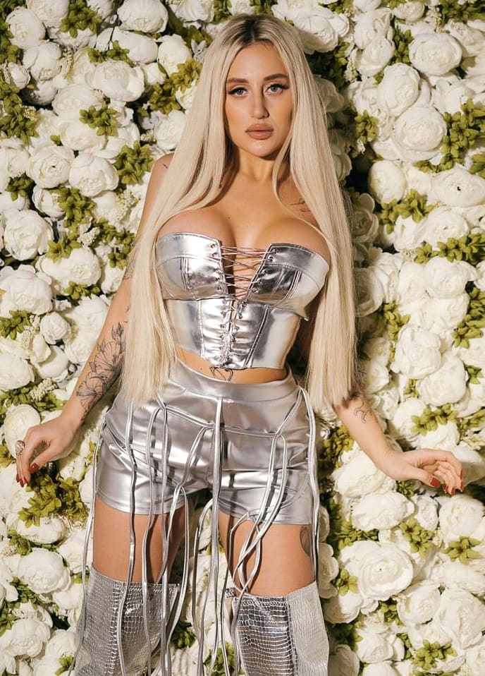 A blonde woman in a silver corset, shorts, and thigh-high boots stands before a wall of white roses, showing arm tattoos.