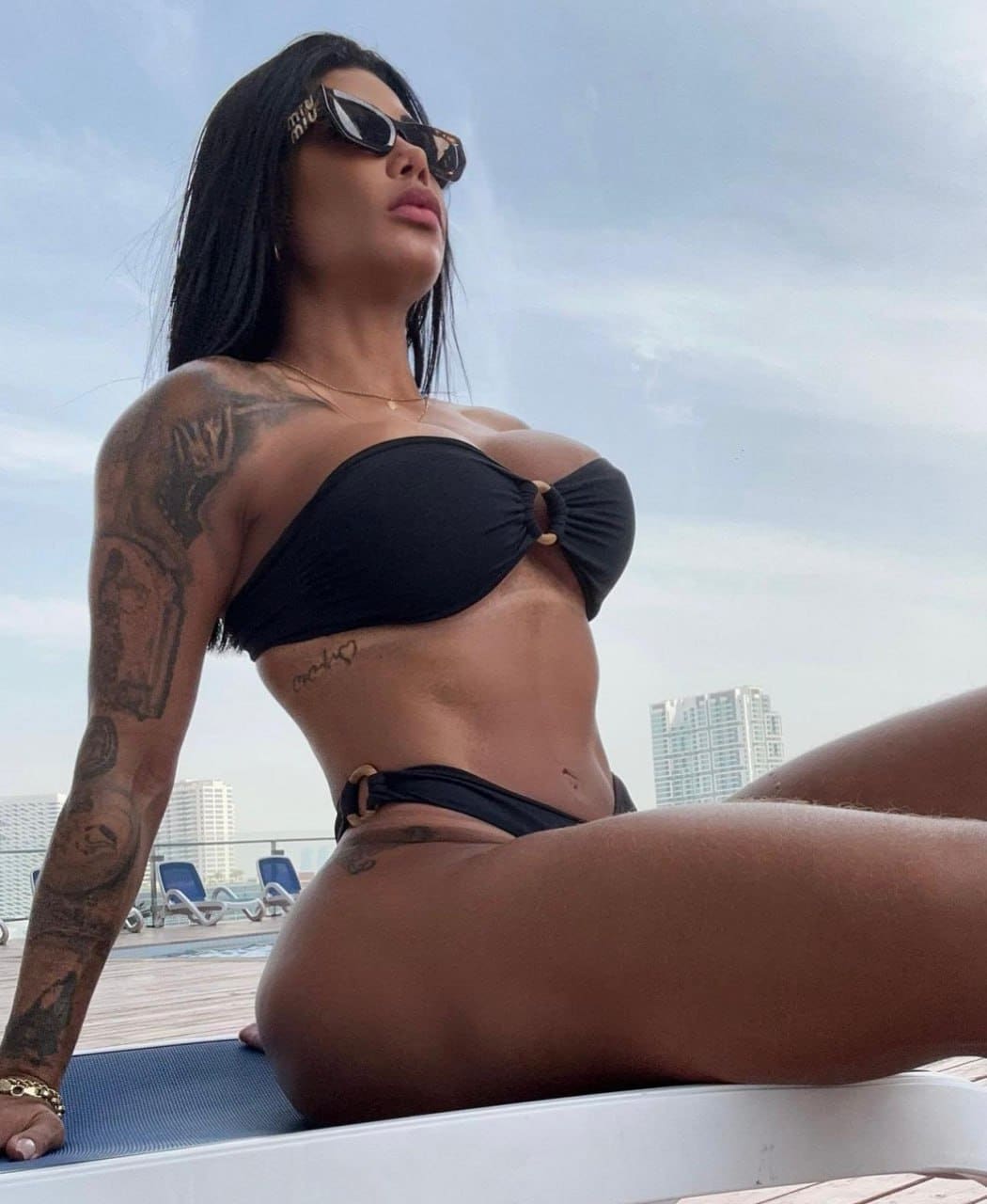 A tattooed woman in a black swimsuit and sunglasses sits on a blue mat by a rooftop pool, with high-rise buildings under a cloudy sky.