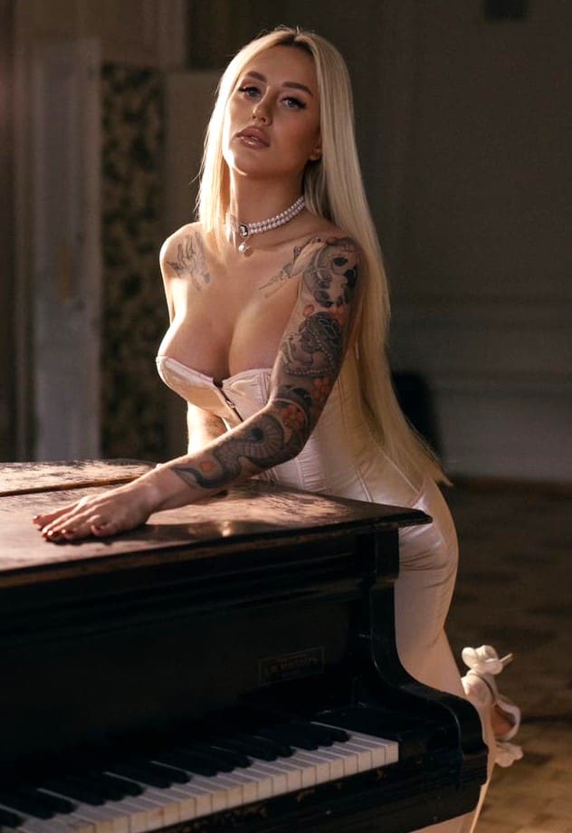 A tattooed blonde in a white strapless dress and pearl necklace leans on an upright piano in a dimly lit room.