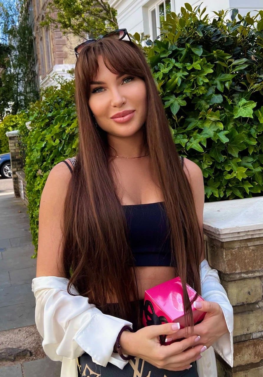 Alyona stands outdoors by a green hedge in a black crop top and draped white shirt, holding a pink box with sunglasses on her head.