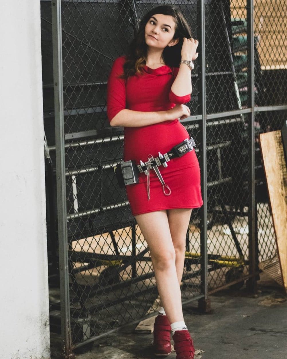 A woman with long dark hair poses by a chain-link fence in a red mini dress with utility belt, white socks, and red lace-up shoes.