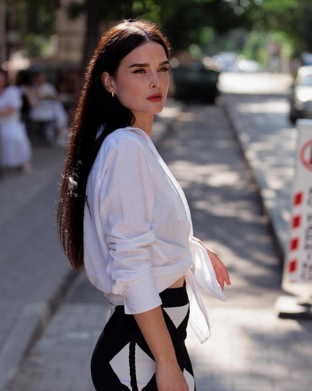 Belle stands on a sunlit city sidewalk in a tied white shirt and geometric skirt, looking back over her shoulder.