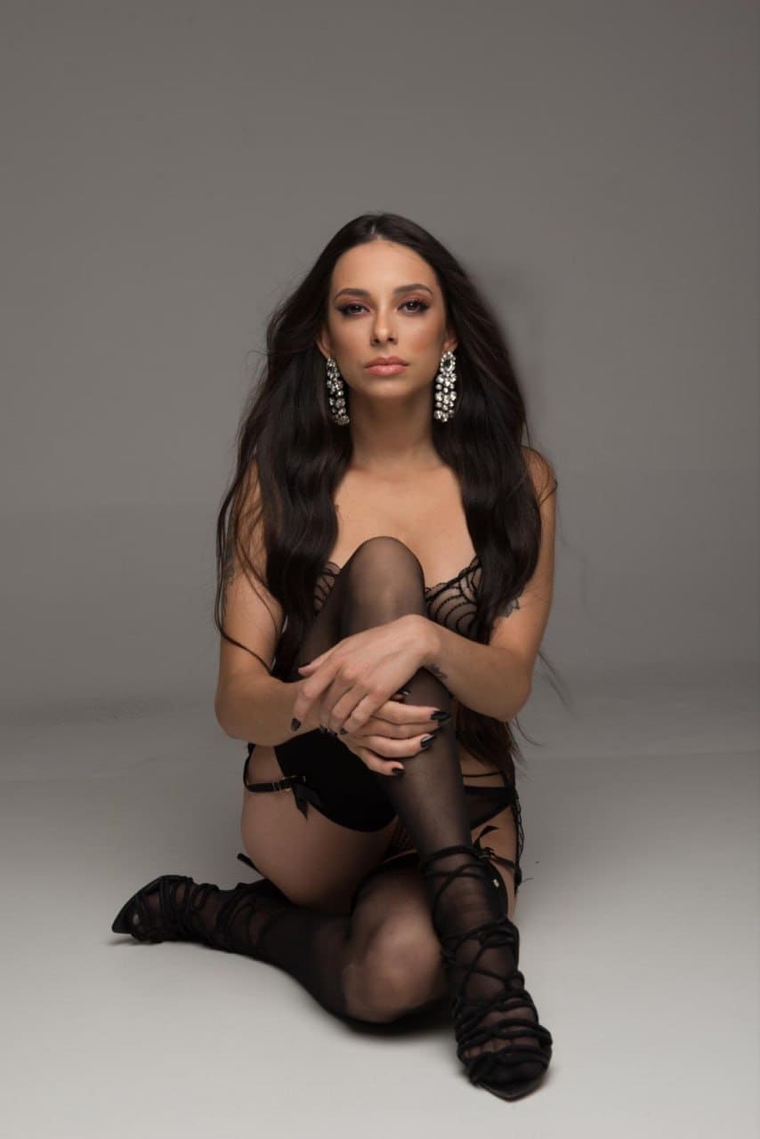 A woman kneels on a light gray studio floor, wearing a black lace lingerie set and stockings, with hands clasped over one knee and large earrings.