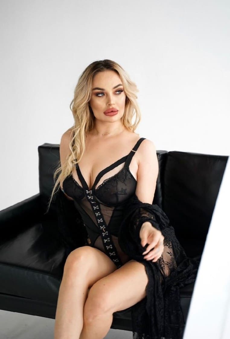 A woman sits on a black leather couch in a black lace bodysuit and sheer robe, set against a plain white background.