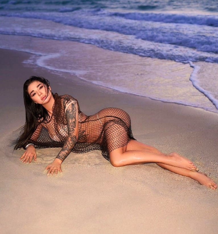 A woman with a tattooed arm lies on wet sand in a sheer black fishnet bodysuit, legs extended, with waves breaking behind.