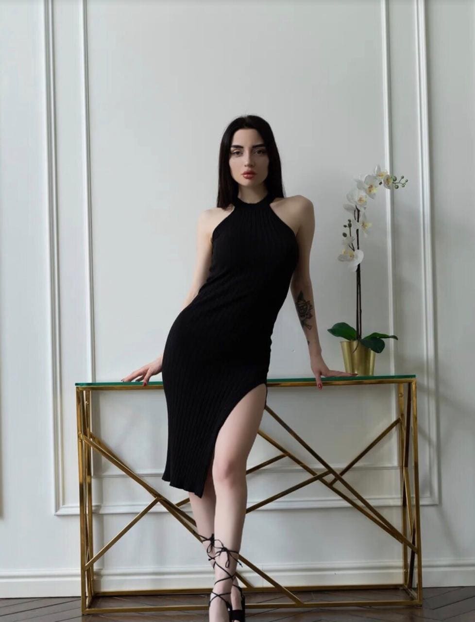 Aleksa stands in a black halter dress with a thigh slit, leaning on a gold console table beside a white orchid in a white paneled room.