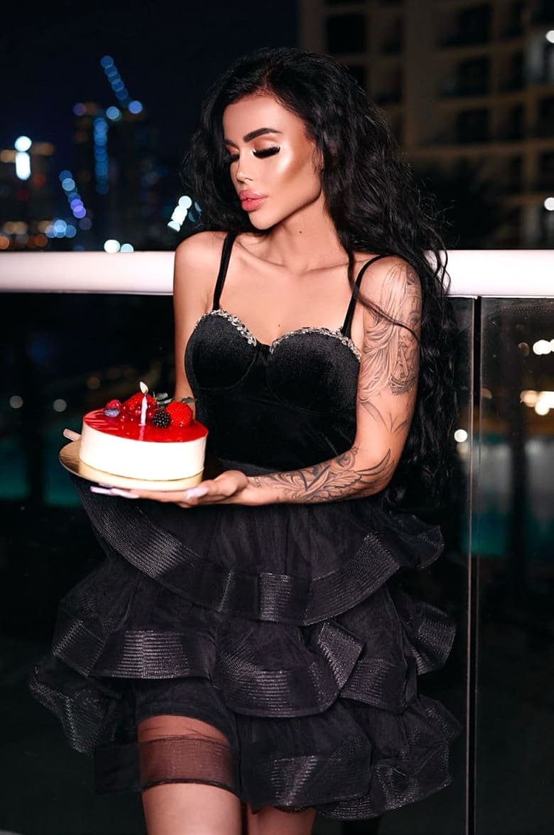 A woman stands on a balcony at night in a black velvet top and ruffled skirt, holding a small cake with a lit candle.