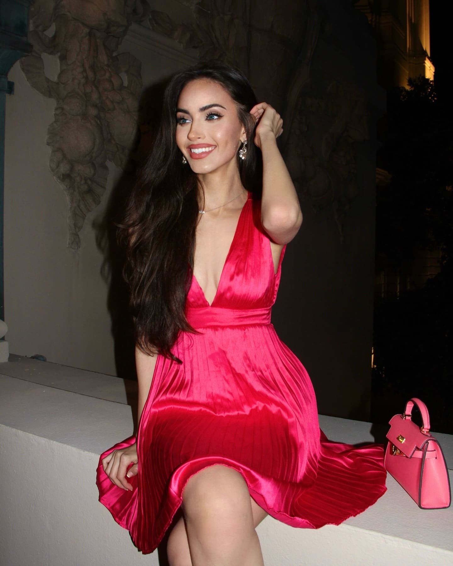 A woman sits on a white ledge in a short deep-V pink satin dress, hand raised to her hair, with a matching pink handbag beside her against a dark ornate wall.