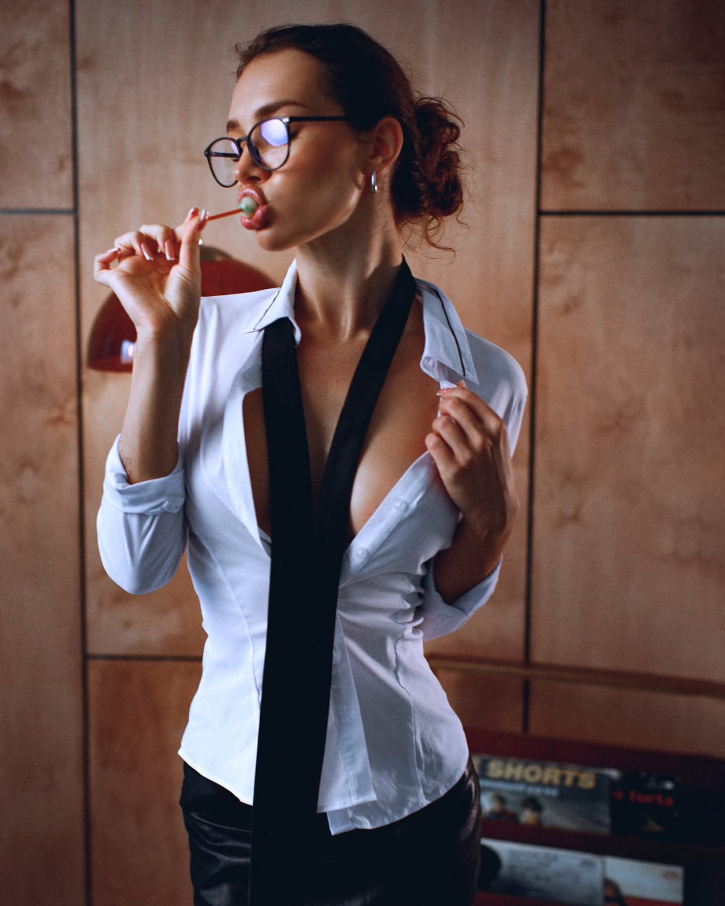 A woman with glasses and a bun holds a lollipop to her mouth, wearing an unbuttoned white shirt and long black tie against wood paneling.