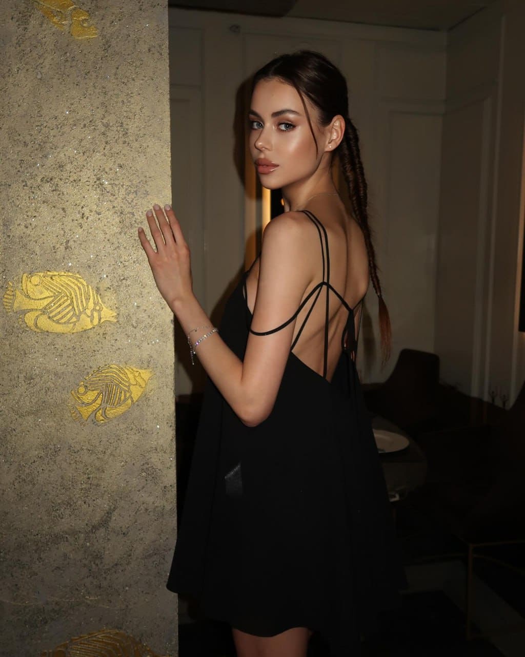 Profile view of a woman in a black dress with crisscross straps, standing against a textured wall decorated with gold fish.