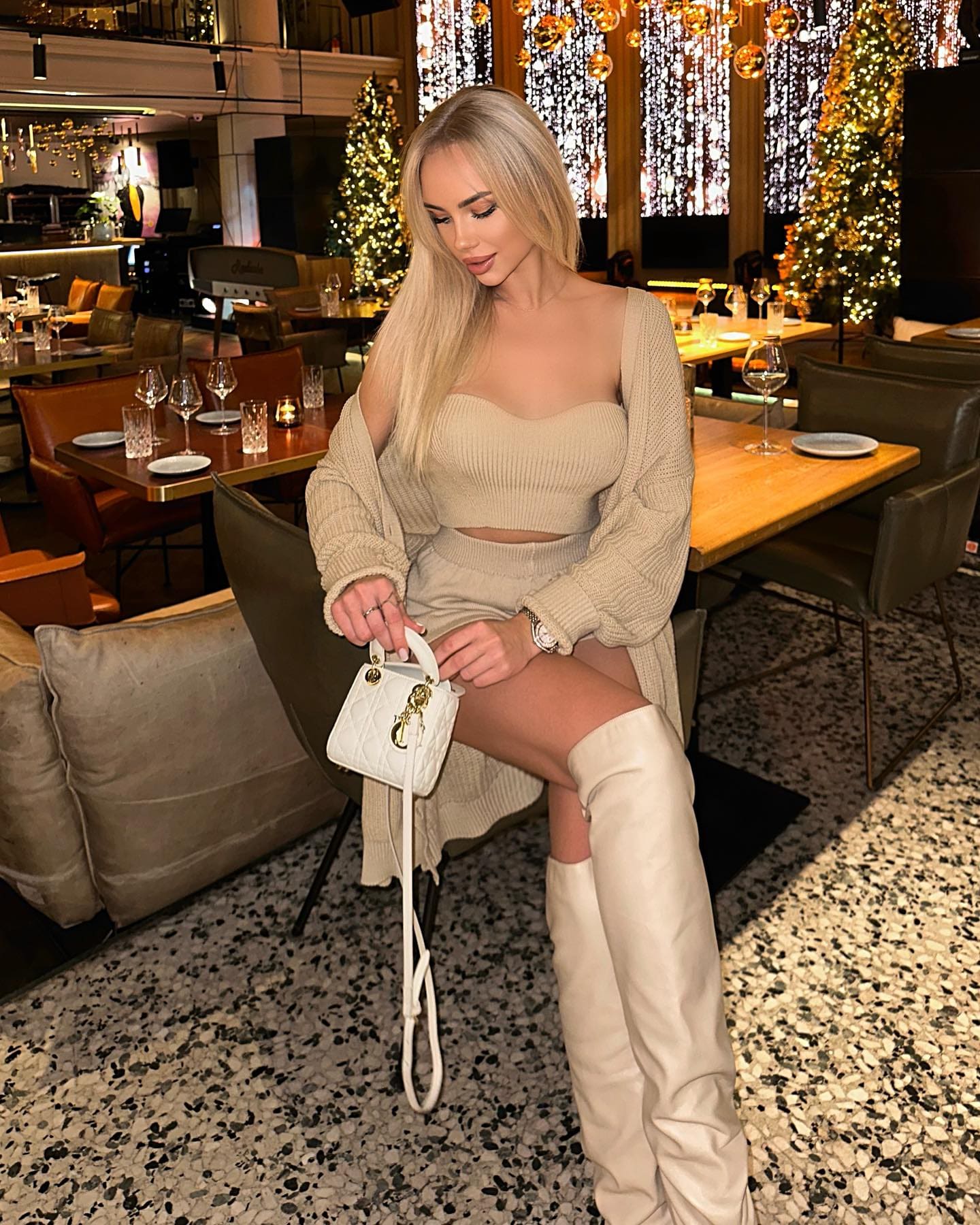 Keli sits in a restaurant with Christmas trees, wearing a beige knit two-piece outfit, white thigh-high boots, and holding a white handbag.