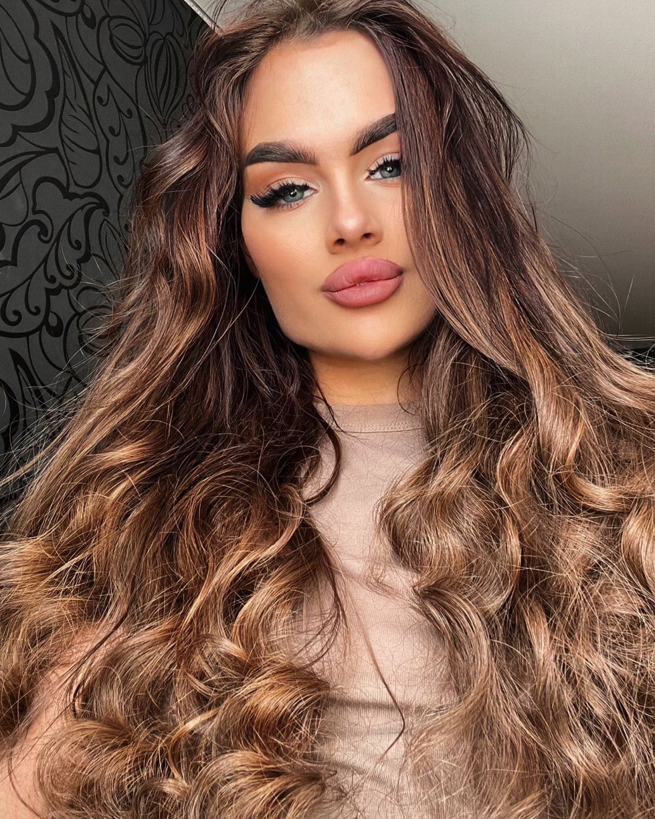 Novella with long wavy brown hair in a light beige top against a dark patterned background.