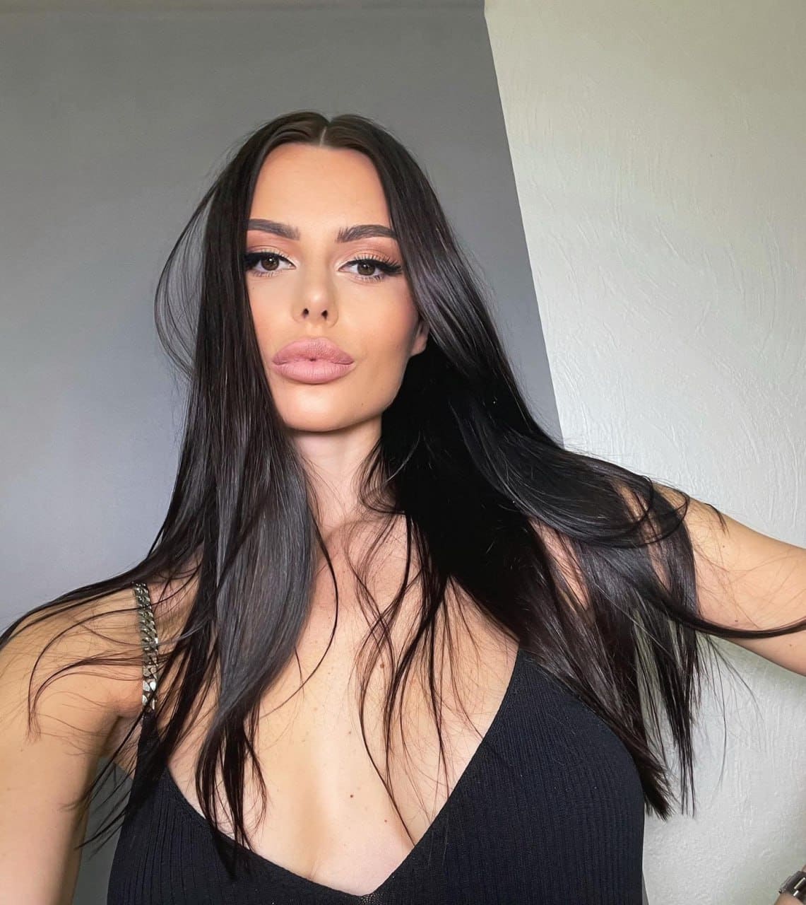Esme stands facing the camera in a black sleeveless top with a deep V-neckline and beaded strap against a plain two-tone wall.