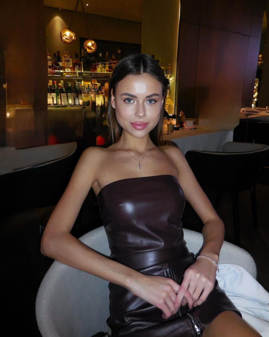 A woman in a strapless dark brown leather dress sits in a light chair, with a bar and shelves of bottles in the background.