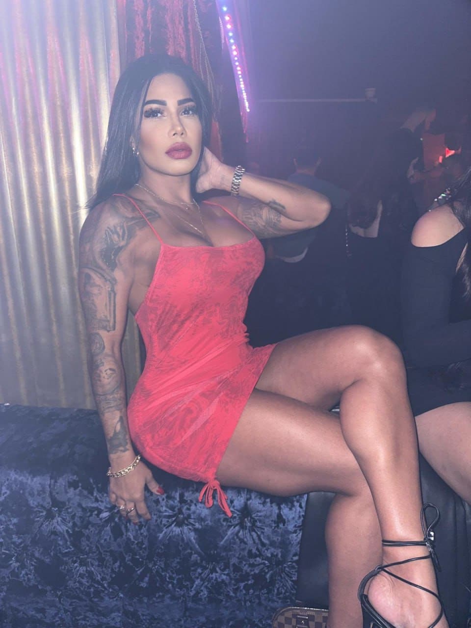 A tattooed woman in a short red lace dress and heels sits on a dark couch, hand to neck, in a dim room with colorful background lights.