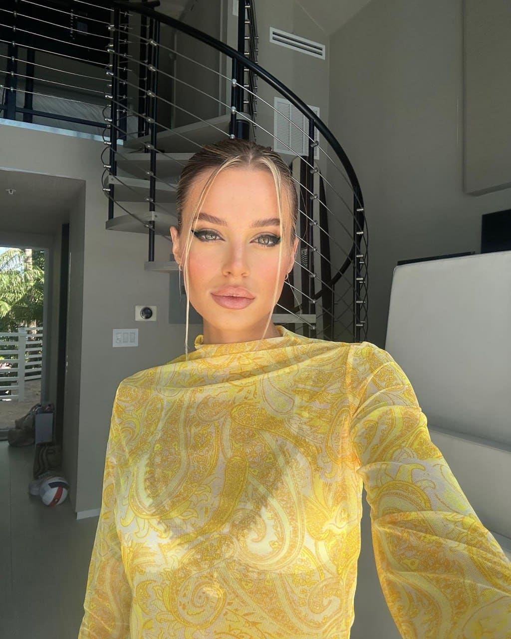 A woman in a sheer yellow paisley long-sleeve top stands in a modern interior with a spiral staircase visible behind her.